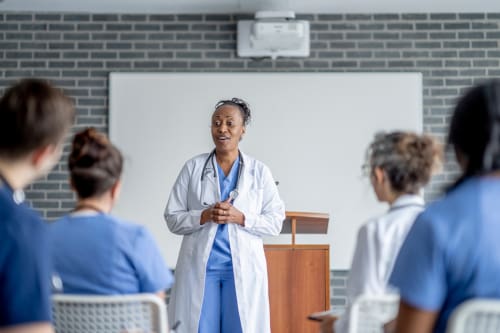 A nursing teacher standing at the front of a classroom with several nurses at desks